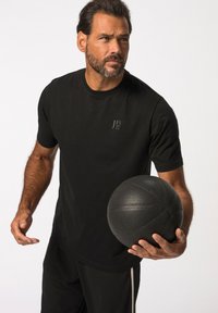 Camiseta deportiva negra con un logo minimalista en el pecho, combinada con pantalones cortos negros. Sosteniendo un balón de baloncesto negro texturizado en un ambiente iluminado.