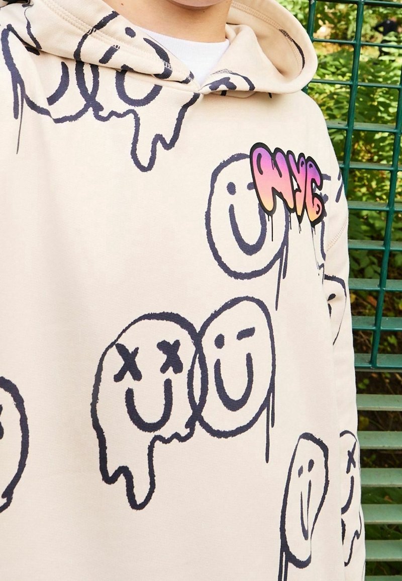 Sweat à capuche beige avec des gribouillis de visages souriants noirs, certains fondant ou rayés, et texte coloré "NYC" sur la poitrine, porté à l'extérieur près d'une clôture verte.