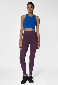 Blå sport-bh med mönster och högmidjade lila leggings, kombinerade med svarta sportskor, mot en enfärgad vit bakgrund.
