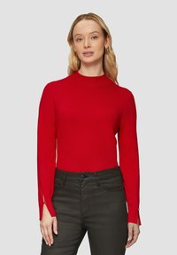Kobieta z długimi blond włosami, ubrana w czerwoną bluzkę z długim rękawem i zapiętymi mankietami oraz ciemne spodnie z wysokim stanem na jasnym tle.
