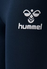 Bílý stylizovaný logo včely s nápisem "hummel" pod ním na tmavě modré texturované tkanině.