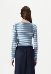 Camisa de manga longa às riscas azuis e brancas com decote redondo, combinada com calças escuras de cintura elástica, vista pela parte de trás.