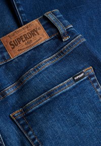 Blå denimjeans med en brun læderlap mærket "SUPERDRY" og orange syninger. En lille sort ekstra mærkat med teksten "SUPERDRY."