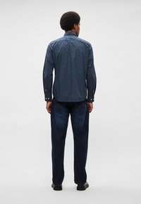 Ναυτικό καρό πουκάμισο με μακριά μανίκια και κουμπωτές μανσέτες, συνδυασμένο με σκούρα μπλε τζιν παντελόνι που διαθέτει ορατές πίσω τσέπες και ίσια γραμμή.