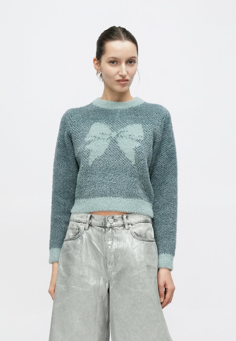 Ανοιχτό μπλε κοντό πουλόβερ από αφράτο νήμα, με μεγάλο σχέδιο φιόγκου σε αντίθετη απόχρωση, συνδυασμένο με γυαλιστερά ασημένια τζιν με φαρδιά μπατζάκια.