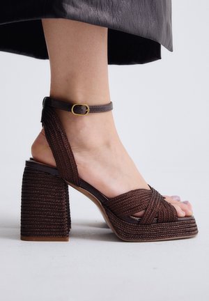 Pie con sandalia de tacón ancho trenzado de color marrón oscuro con correa en el tobillo y hebilla dorada, combinada con una falda negra.