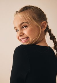 Haut noir à manches longues côtelé avec un col rond. Le sujet a des cheveux châtain clair coiffés en tresses et un léger sourire. Fond neutre.