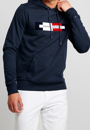 Mężczyzna poprawia kaptur granatowej bluzy Tommy Hilfiger z logo, ubrany w białe spodnie, na jednolitym jasnoszarym tle.