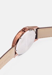 Caixa de relógio em metal cor de rosa com um fundo prateado liso, bracelete de couro castanho e forma redonda. Apresenta uma coroa proeminente de um lado.