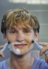 Elektrische trimmer met een grijze behuizing en groene accenten die aan beide zijden wordt vastgehouden. De gebruiker heeft scheerschuim op zijn gezicht en bevindt zich in een badkamer.