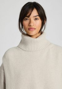 Mujer con cabello oscuro que lleva un suéter de cuello alto acanalado de color crema, mirando hacia adelante con una expresión neutral sobre un fondo liso.