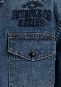Veste en denim de couleur bleue, avec le texte brodé "Petrol & Co Racing", une poche poitrine avec un bouton noir et des détails de couture orange.
