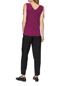 V-hals magenta top met strikdetail op de schouders, gecombineerd met zwarte textuur broek en zwarte instapschoenen. De achterkant heeft een subtiele lage uitsnijding.