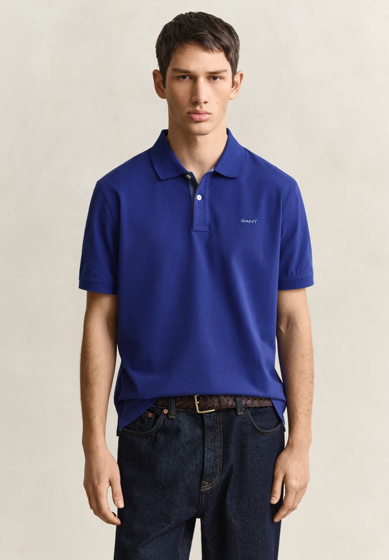 Jeune homme portant un polo bleu à manches courtes avec un petit logo sur la poitrine et un jean foncé sur un fond uni.