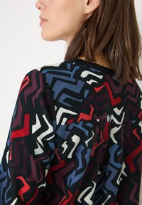 Multifarvet bluse med et geometrisk mønster i rød, blå og hvid på en sort baggrund. Blød stofkvalitet med en afslappet pasform og rund halsudskæring.