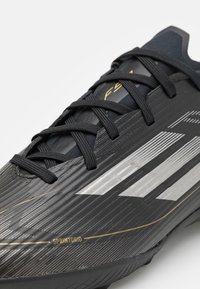 Zapatilla deportiva negra con parte superior sintética texturizada, rayas plateadas, cordones negros y la marca dorada "SPRINTGRID" en el lateral.