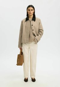 Femme portant une veste beige avec col noir, pull en tricot, pantalon crème, chaussures foncées, tenant un sac à main en daim marron, debout devant un fond blanc.