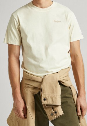 Mężczyzna w kremowym t-shircie z krótkim rękawem, z beżową kurtką zawiązaną wokół talii i ciemnooliwkowymi spodniach.