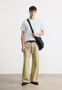 Νεαρός άνδρας όρθιος, φοράει λευκή κοντομάνικη μπλούζα, μπεζ παντελόνι άνετης γραμμής, μαύρη ζώνη, μαύρα παπούτσια και κρατά μαύρη τσάντα χιαστί σε λευκό φόντο.