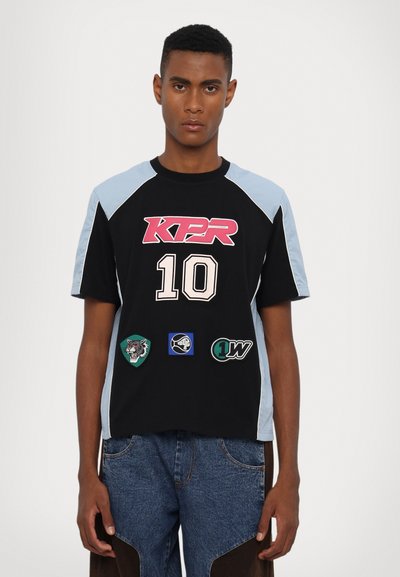 Camiseta de manga corta negra con acentos azul claro, con "KPR" en rosa y "10" en blanco, adornada con parches de varios colores.