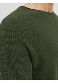 Texturerad grönstickad tröja med runda halsringning och förstärkta sömmar, som visar en närbild av axelområdet.