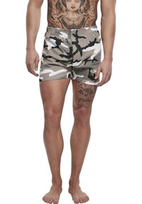 Homme portant un short camouflage gris, blanc et noir, debout pieds nus avec des tatouages visibles sur le torse et la cuisse gauche.