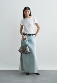 Witte T-shirt, lichtblauwe denim maxi-rok met hoge taille en een voorste split, zwarte riem en een zilverkleurige geweven handtas. Platte zwarte schoenen.