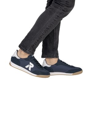 Personne portant des baskets bleu marine et blanches avec des semelles beige clair et un jean noir, debout avec un pied légèrement levé.