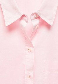 Helles rosa kurzärmliges Hemd mit spitzem Kragen, zwei weißen Knöpfen am Hals und einer Brusttasche auf der linken Seite. Sanfte Textur.