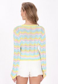 Pull en tricot avec un motif ondulé en bleu clair, jaune et rose. Manches longues et coupe ample. Porté avec un short blanc déchiré.