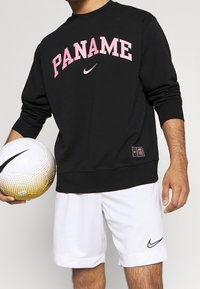Felpa nera Nike con testo rosa "PANAME", abbinata a pantaloni corti bianchi con un piccolo logo Nike nero. Tiene in mano un pallone da calcio texture.