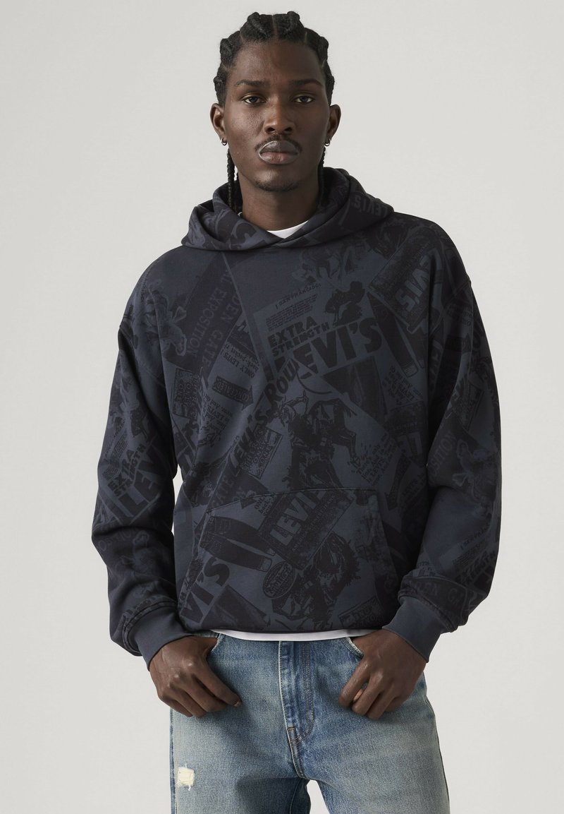 Man with braided hair wears dark patterned Levi's hoodie and light blue distressed jeans, standing against a plain light background.