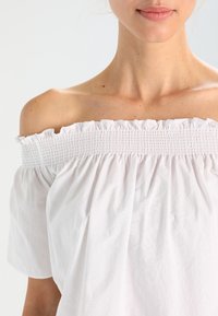 Vit off-shoulder topp i lätt tyg med en smockad halsringning och lösa, flödande ärmar. Slät textur med minimala rynkor.