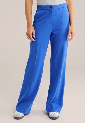 Široké nohavice jasnej modrej farby so zapínaním na gombíky, nosené s bielymi teniskami a svetlosivým topom.