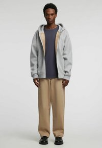 Grå hettejakke med glidelås og beige fôr, brukt over en marineblå skjorte og beige bukser. Antrekket har en avslappet passform og glatt tekstur.