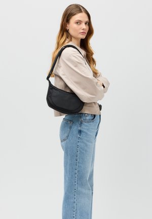 Sort skuldertaske lavet af glat stof, der har en buet form og en bred rem. Personen bærer afslappede blå jeans og en lys skjorte.