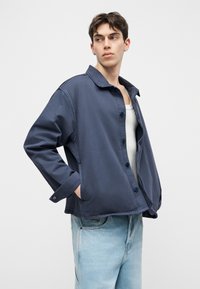 Νεαρός άνδρας που φοράει ένα φαρδύ σκούρο μπλε σακάκι με κουμπιά πάνω από ένα λευκό φανελάκι και ανοιχτόχρωμο μπλε τζιν, κοιτάζοντας προς τα πλάγια με τα χέρια στις τσέπες.