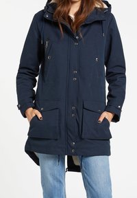 Mujer vestida con una parka con capucha de color azul marino, con bolsillos con solapa, botones a presión, cremallera y cordones, combinada con jeans de un azul claro.