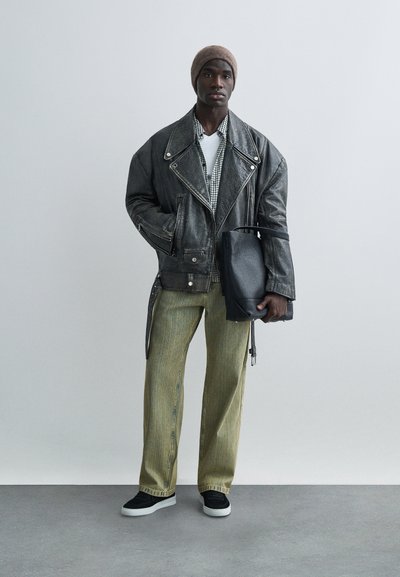 Jaunas vyras, dėvintis rudą kepuraitę, per dydį didelę juodą odinę striukę, žalias kelnes, juodus laisvalaikio batelius, laikantis juodą odinį pirkinių krepšį.