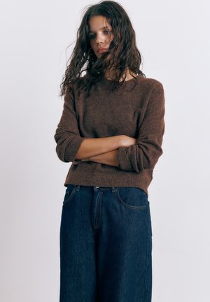 Ung kvinde med langt bølget hår, krydsede arme, iført en brun sweater og mørke viddebukser, foran en neutral baggrund.