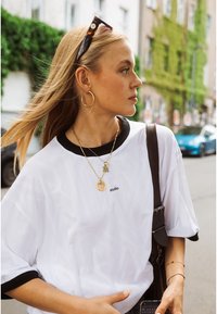 Femme aux longs cheveux blonds portant un t-shirt blanc, des boucles d'oreilles créoles dorées, des colliers superposés, des lunettes de soleil sur la tête et portant un sac à bandoulière noir à l'extérieur.