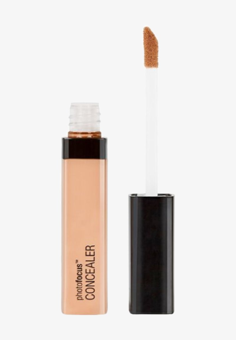 Tube de correcteur liquide beige avec un bouchon noir et un pinceau applicateur incliné levé à côté sur un fond blanc.
