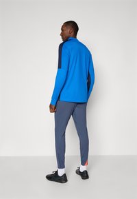 Blå langermet treningstopp med marineblå skulderdetaljer, kombinert med grå smale bukser. Har et sporty design og lett materiale.