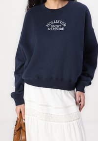 Kvinna som bär en marinblå Hollister Sport & Leisure sweatshirt och en vit kjol med spetsdetaljer, håller en brun läderhandväska.