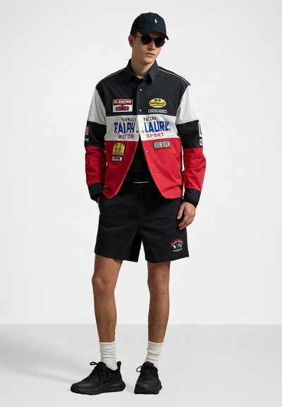 Mehrfarbige Rennjacke mit Aufnähern, mit roten, schwarzen und weißen Abschnitten. Getragen mit dunkelblauen Shorts und schwarzen Turnschuhen. Dunkle Sonnenbrille.