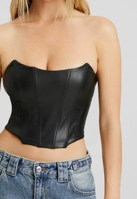 Svart korsett-topp i konstläder med ett hjärtformat urklipp och vertikala sömmar, ihop med hög midja blå denimjeans med knäppning.