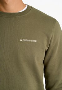 Sudadera verde oliva hecha de un tejido texturizado, con un cuello redondo y un logo bordado en blanco "JUNK de LUXE" en el pecho.