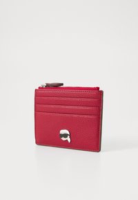 Porta-cartões vermelho em couro com superfície texturizada, apresentando múltiplos slots para cartões e um compartimento com zíper, realçado por um emblema prateado.