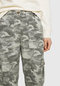 Kamuflażowe spodnie cargo w odcieniach szarości i zieleni, z dużymi bocznymi kieszeniami i zapięciem na guziki. Materiał wydaje się miękki o gładkiej teksturze.