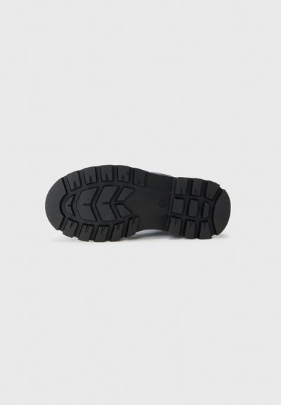 Suela de zapato de goma negra con un patrón de banda de rodadura profundo, que presenta ranuras y agarres circulares para mayor tracción. Superficie duradera y texturizada.